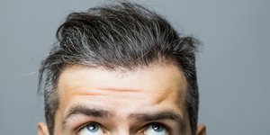 Portrait von einem Mann in mittlerem Alter mit grauen Haare, der seinen Blick fragend nach oben auf das Grau in seinen Haaren richtet