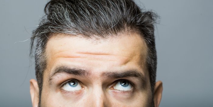 Portrait von einem Mann in mittlerem Alter mit grauen Haaren, der seinen Blick fragend nach oben auf das Grau in seinen Haaren richtet